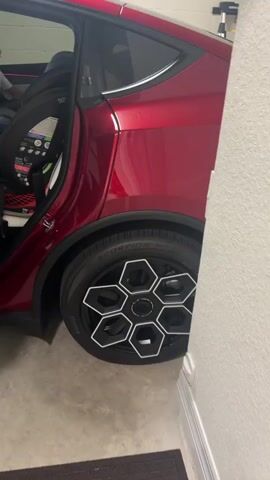 A red car is parked in a garage. The driver's side door is open, revealing the interior of the car. The car has white leather seats and a black steering wheel. There is a large touchscreen display on the dashboard. A person is partially visible in the backseat, wearing a white shirt and blue shorts. The car's wheels have a unique hexagonal design. The garage appears to be well-lit, with the overhead garage door visible in the background. The video pans across the interior of the car, showing the dashboard, steering wheel, and seats. The video also briefly shows the exterior of the car, including the wheel and the rear door.