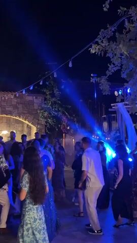 The video shows a crowded outdoor event at night. People are dancing under blue and white lights. The background features an arched stone structure and trees. The atmosphere appears festive and lively, with a DJ booth visible in the distance. The camera pans across the crowd, capturing various individuals and groups enjoying the music and festivities.