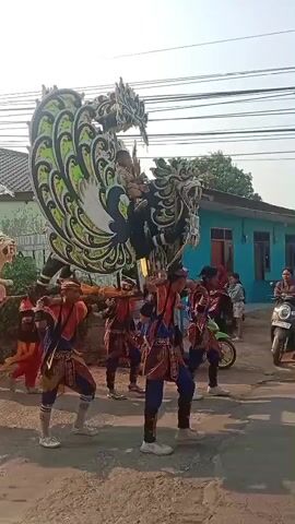 A group of people are participating in a parade. In the foreground, a person on a bicycle rides alongside the procession. To their right, a procession is underway. Several people in traditional costumes, some wearing red pants and ornate tops, are carrying a large, colorful float shaped like a mythical creature. The float has a prominent green and black dragon-like head with a large headdress. Behind the main float, another float resembling a white lion is being carried by people in similar traditional attire. This lion float has a white umbrella above it, and several children are seated on its back. The parade is taking place on a paved street, with houses and buildings visible in the background. There are also power lines running overhead. A few other individuals and a motorcycle are visible further in the background, suggesting a community event. The overall atmosphere appears festive and celebratory.