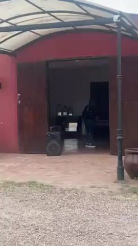 The video shows an outdoor venue. A red building with an open doorway is visible, with a person standing inside the doorway and a speaker outside. The camera pans to show a green lawn with manicured hedges and trees. An old green wagon with red wheels sits on the lawn.  Several tables and chairs are set up under trees in the distance. A white gravel path runs through the lawn, and another old wagon is located on the lawn.