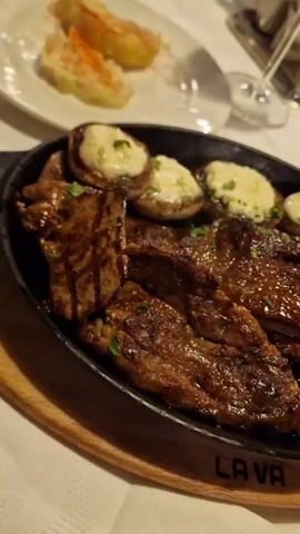 The video shows a sizzling plate of food at a restaurant. The plate is black and round and is on top of a wooden board that says “LAVA”. On the plate are grilled steak strips and mushrooms stuffed with cheese, garnished with parsley. In the background, there’s a white plate with what looks like sliced potatoes or root vegetables, a wine glass, and a few other items on a white tablecloth. The camera slowly rotates around the dish, highlighting the glistening surface of the steak and mushrooms. People are talking in the background. The overall setting suggests a casual dining experience.