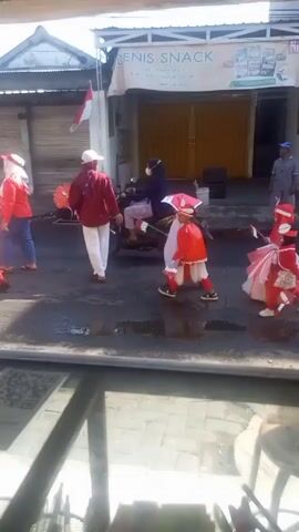A parade marches along a wet street past a building that reads "DENIS SNACK".  Several children dressed in red and white outfits walk in a line, each carrying a small Indonesian flag.  Adults, also in red and white, accompany them.  Motorbikes and other vehicles pass by.  The street surface is damp and reflects sunlight.  An Indonesian flag hangs from the building in the background.