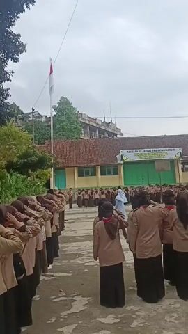 The video shows a group of students in brown uniforms standing in rows, facing a flagpole with the Indonesian flag raised. Some students in the front rows are holding their hands on their foreheads in a salute. In the background, there is a building with a banner that reads "YAYASAN AL-IKHLAS BINA INSAN MANDIRI".  The sky is overcast. The setting appears to be a schoolyard.