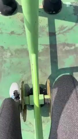 The video shows a person sitting on an outdoor exercise bike and pedaling. The bike is painted light green, and the pedals are weathered with rust. The person is wearing gray pants with a herringbone pattern and white athletic shoes. The surface beneath the bike is painted green and shows signs of wear and cracking. Two black, cylindrical objects are visible in the background. The camera is positioned above, looking down at the person's legs and the bike's pedals.