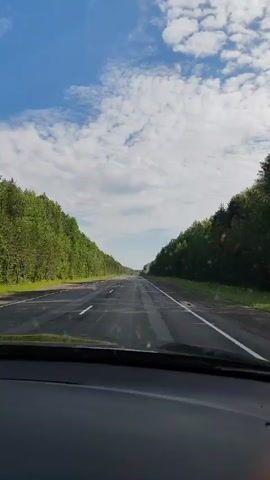 The video shows a view from inside a car driving on a highway. The road is asphalt with white lines marking the lanes. On both sides of the road, there are dense green trees. The sky above is blue with scattered white clouds. The sun is shining. There are other cars visible on the highway in the distance. The perspective is from the dashboard looking forward through the windshield.