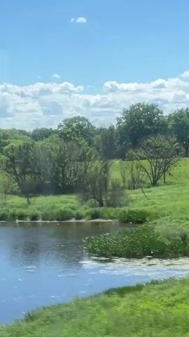 The video captures a scenic view of a body of water with lush green trees and a partly cloudy blue sky. The camera angle suggests it might be from a moving vehicle, possibly a train or car, as there are brief, blurred obstructions that appear to be windows or frames. The water is calm and reflects the sky and surrounding greenery. The environment is natural and appears to be a rural or park-like setting. There are no people or animals clearly visible in the footage. The time stamp indicates the video spans from 00:00 to 00:04.