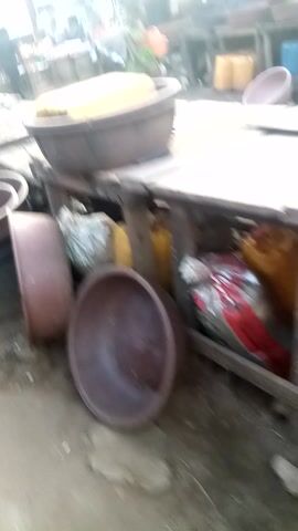 The video provides a brief, handheld glimpse into an active, open-air market in Awka Etiti, Nigeria. The camera is held at an overhead, tilted angle, moving quickly through the space.

**Subjects:**
Numerous people, likely vendors and customers, are visible throughout the market, though individual faces are often obscured by distance or movement. One person in a green and red patterned dress is prominent at a stall. The market features various wooden stalls and tables laden with goods. Large, dark brown basins or bowls are frequently seen, some stacked, others holding items. Yellow plastic containers (possibly jerrycans or similar) are also visible at multiple stalls. Bags of goods, some wrapped in a reflective or foil-like material, are present.

**Activity:**
The primary activity is general market commerce. People are seen moving around, and the person in the patterned dress appears to be attending to goods at a stall. The overall impression is one of a bustling environment.

**Notable Details:**
The scene is brightly lit, suggesting daytime. The market is characterized by simple structures, including corrugated metal roofing and basic building facades in the background. The rapid, often blurry camera movement and tilted angles give a subjective, on-the-ground perspective of navigating a busy market. At approximately 0:02, some dark text is faintly visible on a yellowish wall in the background, though it is too distant and blurred to be clearly deciphered. The market appears to be a central point of activity for the community.