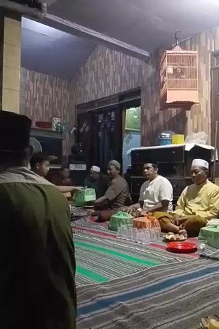 A group of men, some wearing Islamic caps, are gathered in a room. They are sitting on a striped mat on the floor. Some of the men are holding green plastic containers. There are glasses and plates on the mat. In the background, there is a cabinet, a television, and a birdcage with a bird inside. The room appears to be dimly lit.