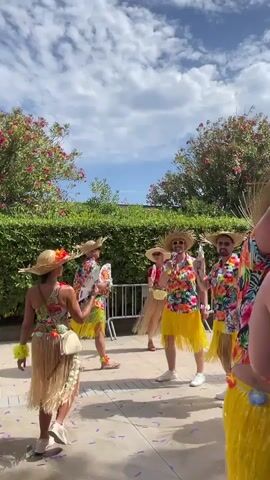 A group of five individuals, appearing to be adults, are dressed in Hawaiian-themed attire, including straw hats, floral shirts, and yellow grass skirts. They are standing outdoors on a paved surface, possibly a patio or walkway. Lush green hedges and trees with pink flowers form a backdrop. The sky above is partly cloudy. The individuals are engaged in a celebratory or festive activity, with some holding what appear to be party favors or decorations, such as leis and possibly confetti or streamers on the ground. The overall atmosphere suggests a party or parade.
