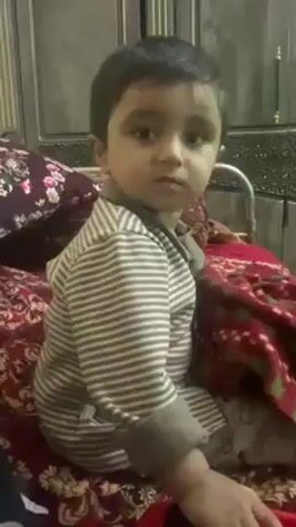 A young child with dark hair, wearing a gray and white striped long-sleeved shirt and matching pants, sits on a bed covered with a maroon patterned blanket and floral pillows. The child is looking directly at the camera with a serious expression. Behind the bed is a dark wooden cabinet or wardrobe. The scene appears to be indoors, possibly a bedroom.