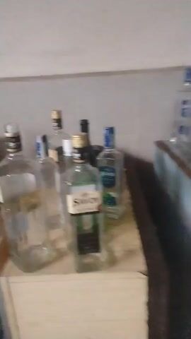 The video shows a close-up of a collection of empty liquor bottles in what appears to be a cluttered storage area. Several bottles are on a wooden surface, with others stored on a shelf below. The bottles are mostly clear glass, with some having blue or black caps and labels that say "Savoy." The environment is dimly lit and appears dusty. The camera moves slightly during the recording.
