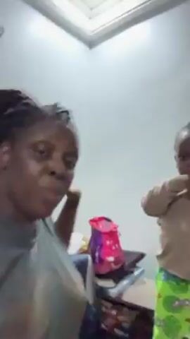 A woman and a young child are standing close to each other, seemingly inside a room. The woman, with dark hair in braids, wears a grey t-shirt. The child, who looks to be about two or three years old, is dressed in a tan shirt and patterned green and yellow shorts. Both are facing the camera, and the child is flapping his hands up and down. A pink and purple backpack is partially visible in the background. The walls are white, and there is a skylight above.