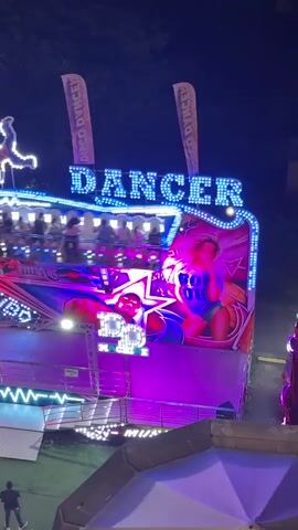 The video shows a carnival ride called "Dancer" at night. The ride is illuminated with colorful flashing lights, primarily blue, green, and pink. The ride itself is a spinning platform with several people on it, experiencing the motion. The background is dark, suggesting it is nighttime. There are also flags with the text "DISCO DANCER" on them visible above the ride. The ride features graphics of people, including women in bikinis and a man wearing sunglasses, with text that includes "MUSIC" and "BOO." There are also stars and abstract patterns as part of the ride's decoration. The overall impression is a vibrant and exciting atmosphere typical of a carnival or amusement park.