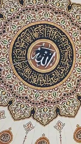 A stationary, low-angle shot shows a beautifully decorated ceiling inside a mosque, adorned with intricate geometric patterns and Islamic calligraphy. At the center is a large circular design with "Allah" written in Arabic script in white on a brown background, surrounded by more calligraphy in black. The outer part of the circle features a complex pattern of intertwining floral and geometric shapes in shades of brown, gold, black, and white. There is a small metal object hanging from the center of the ceiling, possibly a light fixture or a decorative element. The background is a lighter beige color, also with decorative patterns.