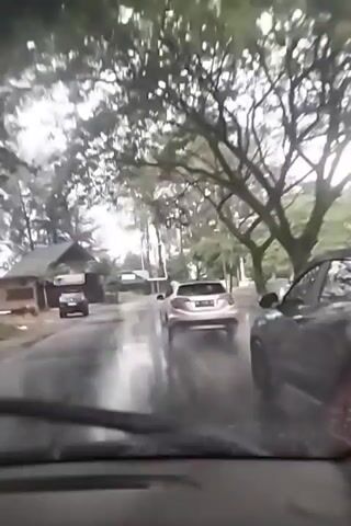 This video shows a view from inside a car traveling on a wet road during what appears to be a rainy day. The road is lined with trees and some buildings are visible in the background, suggesting a semi-urban or suburban setting. Several cars are on the road, including a white SUV in front of the filming vehicle and a darker SUV further ahead. A bus can also be seen further down the road. The weather is overcast, and the road surface is slick with water, indicating rain. The time of day is unclear, but the lighting suggests it could be daytime. There is no visible text in the video. The scene captures a common experience of driving in adverse weather conditions.