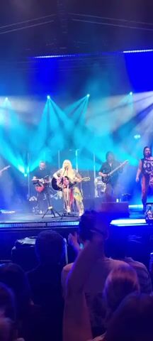 A live band is performing on stage under blue lights and smoke machines. A female singer with blonde hair is center stage, playing an acoustic guitar and singing. She is wearing a brightly colored, patterned outfit. To her left, a male musician plays a guitar, and to her right, two male musicians play guitars. Behind them, a drummer is visible. The audience is visible in the foreground, with several people holding up phones to record the performance. The stage appears to be elevated.