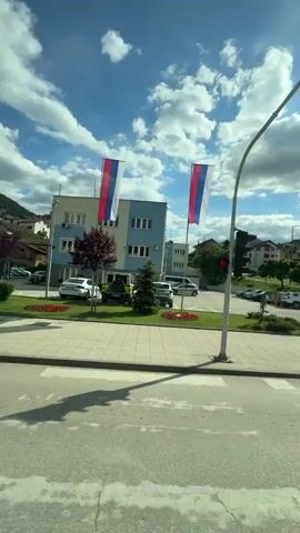 The video depicts a scene from inside a moving vehicle as it passes a building with a light blue and white facade. Two flags with a red, blue, and white horizontal stripe design are visible on poles near the building. The building appears to be some sort of public or government building. There are cars parked in front of the building and in a nearby parking lot. Across the street, there is a small, well-maintained public park with flowers, bushes, and a few small trees. The sky is blue with scattered white clouds.