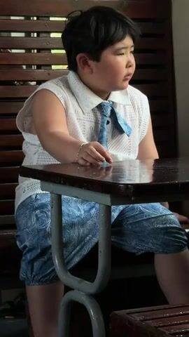 A boy sits at a table inside a train. He is wearing a white shirt with a blue tie, and blue patterned pants. He has short, dark hair. The table is black and reflective. The train has a wooden bench with a metal frame. People can be heard speaking in the background.