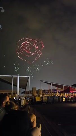 A drone light show displays a large, illuminated rose in the night sky. The rose is primarily red and pink, with green lights forming its stem and leaves. Below the sky show, a group of people are seated on wooden benches, facing away from the camera and towards the spectacle. In the background, there are structures with white canopies and lights, suggesting an outdoor event or venue. The overall scene depicts an evening outdoor entertainment event.