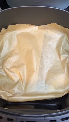 A person places a large yellow tortilla in an air fryer. The air fryer is lined with parchment paper. The tortilla is round and has some brown spots. A hand is visible placing the tortilla. There is also background noise.