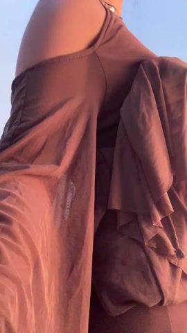 The video shows a woman wearing a brown dress with an open back, adorned with dangling gold chains and sequins. The dress has long, flowing sleeves. Her hair is styled in a bun. She turns slightly to her left. The background is a plain, white wall.