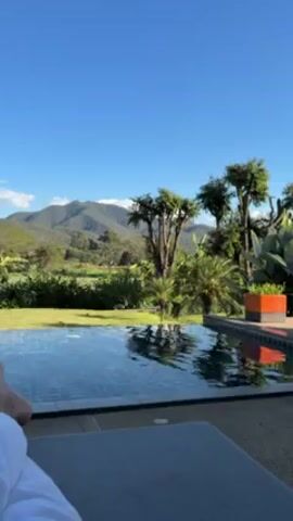 The video shows a person relaxing by an outdoor infinity pool on a sunny day. The person is lying on a lounger, with their feet visible towards the camera. Beyond the pool is a green lawn, trees, and a mountain range in the distance under a clear blue sky. The pool has dark blue tiles and the water surface is slightly rippled. There is a plant in an orange pot on the right side of the pool.