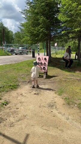 A young child wearing a checkered outfit and a hat stands on a dirt path, facing a pink game board with multiple circular targets. The child is reaching towards the game board, possibly about to play. The game board is set up outdoors, with trees and grass surrounding it. In the background, there is a road with cars, a barrier, and a few adults sitting on a bench under a tree. The sky is partly cloudy.