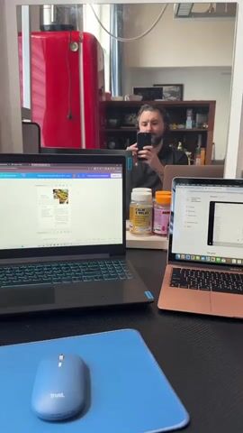 The image captures a home office setup with two laptops and a computer mouse on a dark desk. A person, presumably the photographer, is visible in the reflection of a mirror positioned above the desk, holding a smartphone to take the picture.

On the desk, to the left, is a black laptop with a backlit keyboard displaying what appears to be a document with an image of food on a white background, possibly in a design application like Canva (indicated by the logo). To the right, a rose gold/pink Apple MacBook is open, showing text on its screen. In front of the black laptop, a blue wireless mouse with "Trust." written on it rests on a blue mouse pad. Between the two laptops, two supplement bottles are visible: a white one labeled "CALM & RELAX" and "MAGNESIUM," and an orange one with "SKIN" printed on it.

The mirror's reflection shows a red refrigerator behind the person, and shelves with various items further in the background, suggesting a kitchen or living area. The person in the reflection has dark hair and a beard, wearing a dark shirt. The overall setting is indoors. The audio clip features the word "Работаем" (Rabotaem), which translates to "We are working" or "I am working."