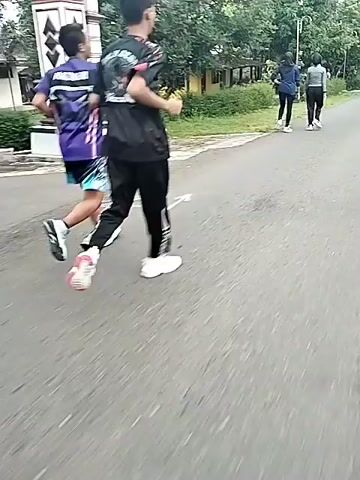 Two people are walking on a paved road, dressed in casual athletic wear. The camera follows from behind as they walk, soon passing more people doing the same. The road has a painted center line and the area is lined with grass and trees, with houses visible between the trees.