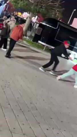 At night, two people, presumably male and a child, are celebrating outdoors, likely at a public event. The man, wearing a black jacket and pants, and the child, in a red jacket and light blue pants, are running in circles, excitedly. The child wears a red hat with green accents, suggesting a specific national affiliation or team support. A large red sign with white lettering, associated with a sporting event, stands nearby. In the background, a crowd of people is visible, suggesting a festive atmosphere. A black trailer is parked near the sign. The ground is paved with a tile-like pattern.