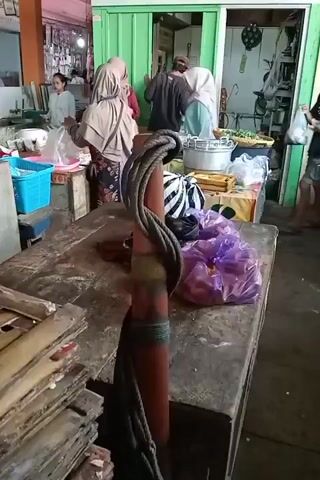 The video shows a bustling market scene with several people present. In the foreground, a thick rope is wrapped around a reddish-brown pole. Behind the pole, a wooden table is visible, covered with various items including a purple plastic bag filled with red items, a striped fabric, and other unidentifiable objects. Further back, a green structure, possibly a stall or shop, is open, revealing an interior with more people and goods. On the left side of the frame, a woman in a light blue hijab is attending to a display of items, with a blue plastic basket nearby. Other individuals are seen interacting, some appearing to be vendors and customers. One person, a young child in a pink and white dress, stands near the right edge of the frame. The overall environment appears to be an outdoor or semi-outdoor market.