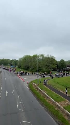 The video shows a group of people gathered near a road, likely participating in a walk or march. The road has traffic cones directing movement and white arrows painted on the surface. Trees and green spaces surround the road. Some people are walking on a paved path alongside the grass. The sky is overcast.