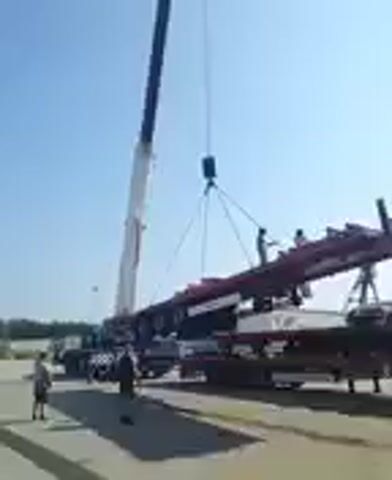 This media file shows a crane lifting a large, red, rectangular object that appears to be a modular building component or a shipping container frame. The object is being lowered onto a flatbed trailer that is already carrying a similar, purple object.

There are several people visible, some standing on the ground and at least two on top of the red object, seemingly guiding its placement. The crane's arm is tall and white with black accents, and it is extended over the scene. The sky is clear and blue, suggesting it is daytime. The ground is paved and appears to be an outdoor industrial or storage area. There are other vehicles and structures in the background, including what looks like other trailers and possibly a building.

The activity depicted is the loading or unloading of heavy equipment or materials using a crane. The overall mood seems to be one of focused work, with no clear indication of positive or negative emotions. The location is identified as Houthalen, Belgium. No visible text is present in the media file.