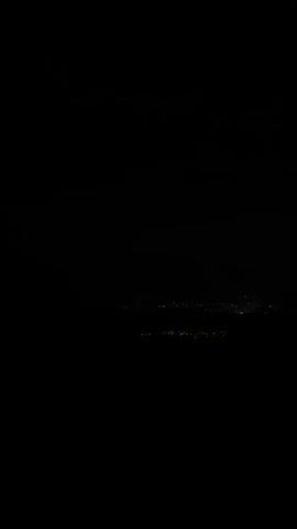 The video shows a night view from a vehicle. Streetlights with white lights are visible in the distance. The road has a yellow line on the right side. There are also blurry lights in the far distance, possibly indicating a city or town. The sky is dark and appears to be clear. The overall scene is dark and somewhat abstract due to the motion of the vehicle and the low light conditions.