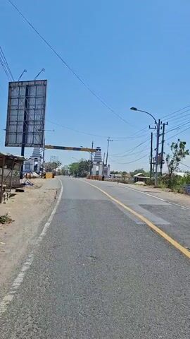 The video begins with a view of a road leading towards a large archway. The sky is clear and blue. On the left side of the road, there is a large billboard. The archway is white with decorative elements and a banner across the top. Orange cones are visible on the sides of the road near the archway. As the camera moves forward, the road curves slightly to the right. The scenery changes to a view of the sea. The water is a light blue-green color, and there is a shoreline with sand and some debris. A low wall or structure is visible along the edge of the water. Power lines are visible against the sky throughout the video.
