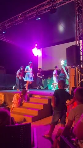A group of children and adults are dancing on a stage lit by purple lights. A few children are seated in front of the stage watching the performance.