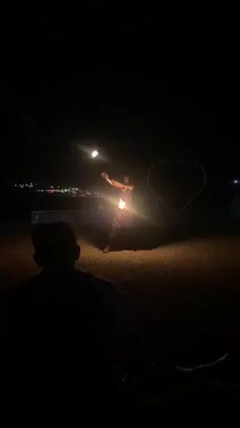 A person is seen performing a fire-breathing act at night. They are spraying a large burst of fire from their mouth. A large, illuminated heart shape is visible in the background, along with some distant lights that suggest a settlement. The ground appears to be dry and dusty. In the foreground, two individuals are silhouetted, facing away from the camera and towards the performer, suggesting they are spectators. The lighting is dim, with the fire being the primary light source in the immediate vicinity of the performer. The scene appears to be outdoors, possibly in a desert or a rural area.