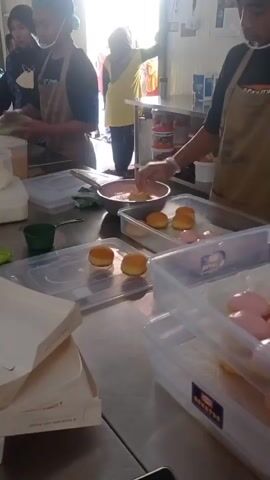 The video shows a group of people in what appears to be a kitchen or food preparation area. In the foreground, a person wearing an apron and gloves is dipping a small, round pastry into a bowl of pink frosting or glaze. To the left of this person, another individual in an apron is working with food items on a counter. In the background, other people are visible, including one wearing a yellow shirt and headscarf standing near a doorway. The environment includes stainless steel counters, plastic containers, and what looks like packaged goods on shelves. The overall scene suggests food production or packaging.