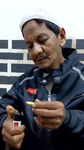 A man in a black jacket and a white knitted cap is lighting a cigarette. He is using a red and white lighter. The background is a white wall with black brick-like patterns.