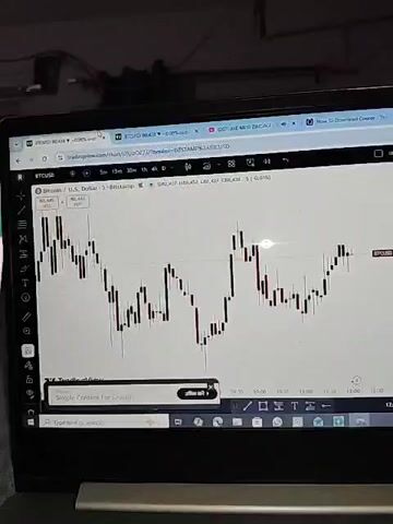 The video shows a laptop screen displaying a trading chart, likely for Bitcoin (BTCUSD), as indicated by the title bar. The chart shows price fluctuations over time, with candlesticks representing price movements. There are also various trading tools and indicators visible on the screen. The environment appears to be dimly lit, suggesting it might be indoors, possibly at night. No people are visible in the video, but the presence of a laptop suggests someone is using it. The overall scene is focused on financial data visualization, characteristic of online trading platforms.