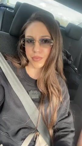 A woman with long brown hair, wearing a black sweatshirt, gold-rimmed sunglasses, and a seatbelt, sits in the back of a car. She looks directly at the camera and smiles. A black backpack and a jacket are visible on the seat behind her. The car is moving, and music plays in the background.