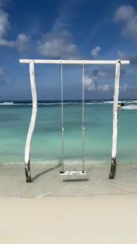 A stationary shot shows a wooden swing set on a sandy beach. The swing seat is suspended by two ropes from a horizontal bar supported by two weathered, white-painted posts. The swing is positioned at the edge of the turquoise ocean, where small waves are breaking. In the background, a person on a jet ski is visible in the distance, creating a wake on the water. The sky above is blue with scattered white clouds.