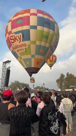 A large crowd of people gathers outdoors, many pointing or filming with their phones to the sky filled with hot air balloons. A prominent hot air balloon with multicolored square patterns and the words "Sky Balloons MEXICO" is visible in the foreground. Behind the crowd, there are stage towers with speakers. In the distance, several hot air balloons drift across a partly cloudy sky, with a pyramid-like structure visible on the horizon. The overall atmosphere suggests a festival or event centered around hot air ballooning.