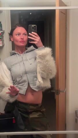 A woman stands in front of a mirror taking a selfie. She is wearing a gray zip-up hoodie with light-colored fur cuffs and camouflage pants. The hoodie is partially unzipped, revealing her midriff. She initially poses with an open hand gesture and then touches her stomach. The background shows a bathroom with a door and some items on a shelf. The mirror reflects a dimly lit hallway or room.
