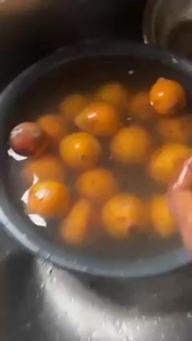 The video shows a black plastic basin filled with water and yellow, plum-sized fruits soaking in it. The basin sits inside a stainless steel sink. A hand with dark skin moves the fruit around in the water. Some of the fruits have dark spots or blemishes on their surface. The water appears slightly murky. The camera zooms in and out, focusing on the fruit in the water.