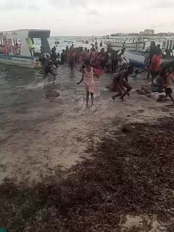 The video shows a crowded beach with many people, mostly children, playing in the water. The water appears murky, and there is a significant amount of seaweed or other organic material washed up on the shore. Several small boats are anchored near the shore. The sky is overcast. People are standing, splashing, crawling, and generally interacting in the water and on the beach. Buildings can be seen in the background.