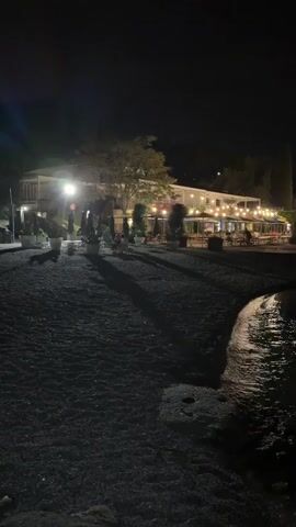 The video shows a nighttime view of a beachside restaurant or bar. The building is illuminated with numerous string lights and interior lights, creating a warm glow. Tables and chairs are set up on an outdoor patio area, and a few people can be seen in the distance, likely patrons or staff. The foreground is a dark, pebbled beach that slopes down to the water's edge. Gentle waves are lapping at the shore, reflecting the lights from the building. Trees and some potted plants are visible around the establishment. The overall ambiance is relaxed and inviting.