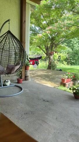 A young woman with long blonde hair sits at a wooden table, smoking a cigarette and looking at her phone. Beside her on the table are two glasses and a black purse. Behind her, a hanging egg chair is visible, with a soccer ball at its base. The scene appears to be an outdoor patio or porch, with greenery and a tree in the background. There are also clothes hanging on a clothesline in the distance.