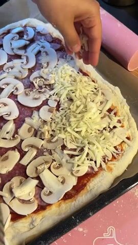 A person is making a pizza. The pizza dough is rectangular and is on a baking sheet lined with parchment paper. The pizza is topped with tomato sauce, salami, sliced mushrooms, and shredded cheese. The person is adding more shredded cheese to the pizza with their hands. A pink rolling pin is visible in the background.