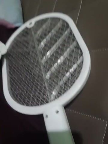 The video begins with a close-up of a white electric mosquito swatter. The camera then pans to the left, revealing a bed with several stuffed animals and pillows. One stuffed animal is yellow and resembles a chick, lying on a pink pillow with cartoon characters. To the left of the yellow stuffed animal are several other stuffed animals, appearing to be white and blue. A dark blue pillow or blanket is also visible on the bed. A small white object with text is lying on the bed in front of the pink pillow. The camera continues to pan, showing more of the bed and stuffed animals, and then moves towards a corner where a brown textured wall is visible. The room appears to have green walls. The lighting is dim. The video ends with a brief glimpse of the same electric mosquito swatter.